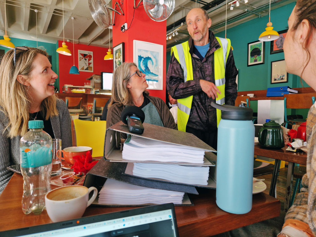 The photo shows a table in a busy coffee shop. Its surface is covered in large legal files and folders, coffee cups and water bottles. The PILS and SWOT teams are huddled around, chatting and smiling.